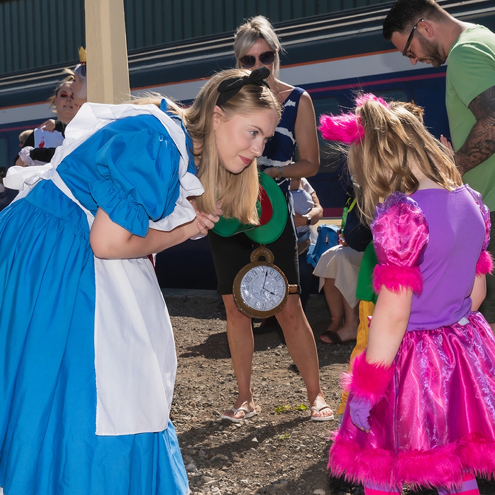 Picture of actor dressed as Alice from Alice in Wonderland bent down talking to a young girl dressed as the cheshire cat on the train platform during The Mad Hatter's Travelling Tea Party